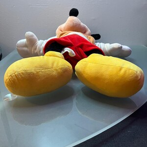 May include: A plush Mickey Mouse doll, lying on its back, features a red shorts, white gloves, and large yellow shoes. The doll has a black head and ears, with a tan face and a black nose. The doll is on a reflective surface.