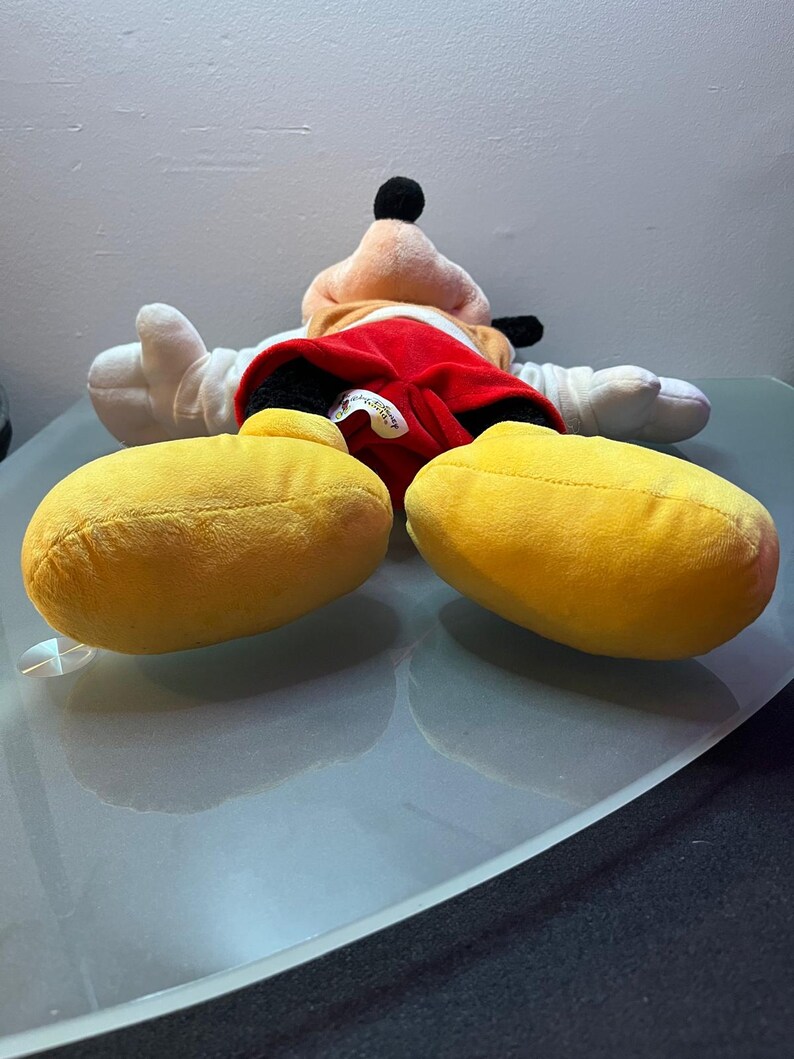 May include: A plush Mickey Mouse doll, lying on its back, features a red shorts, white gloves, and large yellow shoes. The doll has a black head and ears, with a tan face and a black nose. The doll is on a reflective surface.