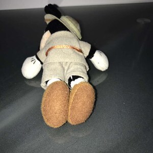 May include: A small plush toy of a cartoon mouse wearing a safari outfit, including a khaki shirt and shorts, a tan hat, and brown shoes. The toy has white gloves and black ears. The mouse is lying on its stomach.