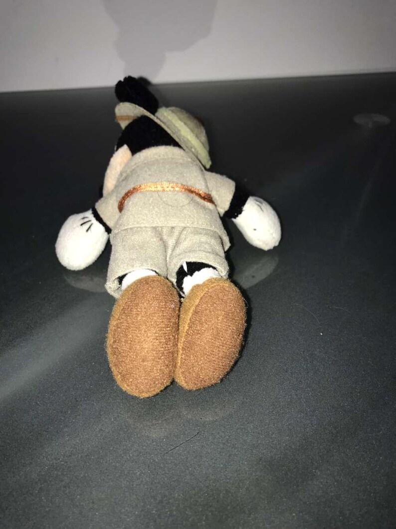 May include: A small plush toy of a cartoon mouse wearing a safari outfit, including a khaki shirt and shorts, a tan hat, and brown shoes. The toy has white gloves and black ears. The mouse is lying on its stomach.