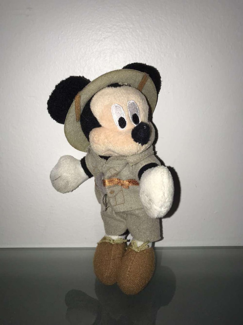 May include: A plush Mickey Mouse toy dressed in a safari outfit, including a khaki-colored hat, jacket, and shorts. The toy has black ears, a black nose, and white gloves and feet. The outfit has gold-colored accents.