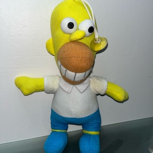 May include: A soft toy of Homer Simpson, with a yellow head, large black and white eyes, and an orange mouth. He is dressed in a white shirt, blue trousers, and blue shoes. A white string is attached to the top of his head.