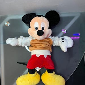 May include: A plush Mickey Mouse toy, featuring black ears, a tan face, and a black nose. Mickey wears a tan shirt, red shorts, white gloves, and yellow shoes. The toy is a classic Disney character.