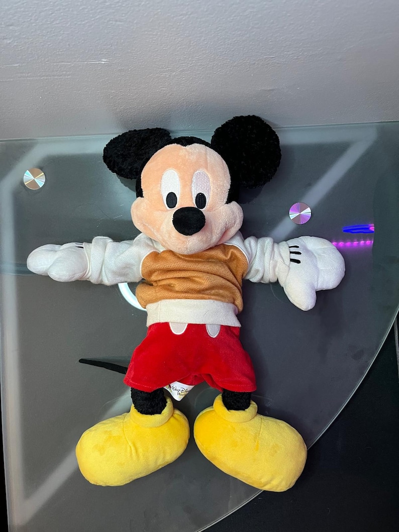 May include: A plush Mickey Mouse toy, featuring black ears, a tan face, and a black nose. Mickey wears a tan shirt, red shorts, white gloves, and yellow shoes. The toy is a classic Disney character.