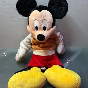 May include: A plush Mickey Mouse doll with black ears, a tan face, and a black nose. He wears a tan and orange shirt, red shorts, and large yellow shoes. The doll is a classic Disney character.