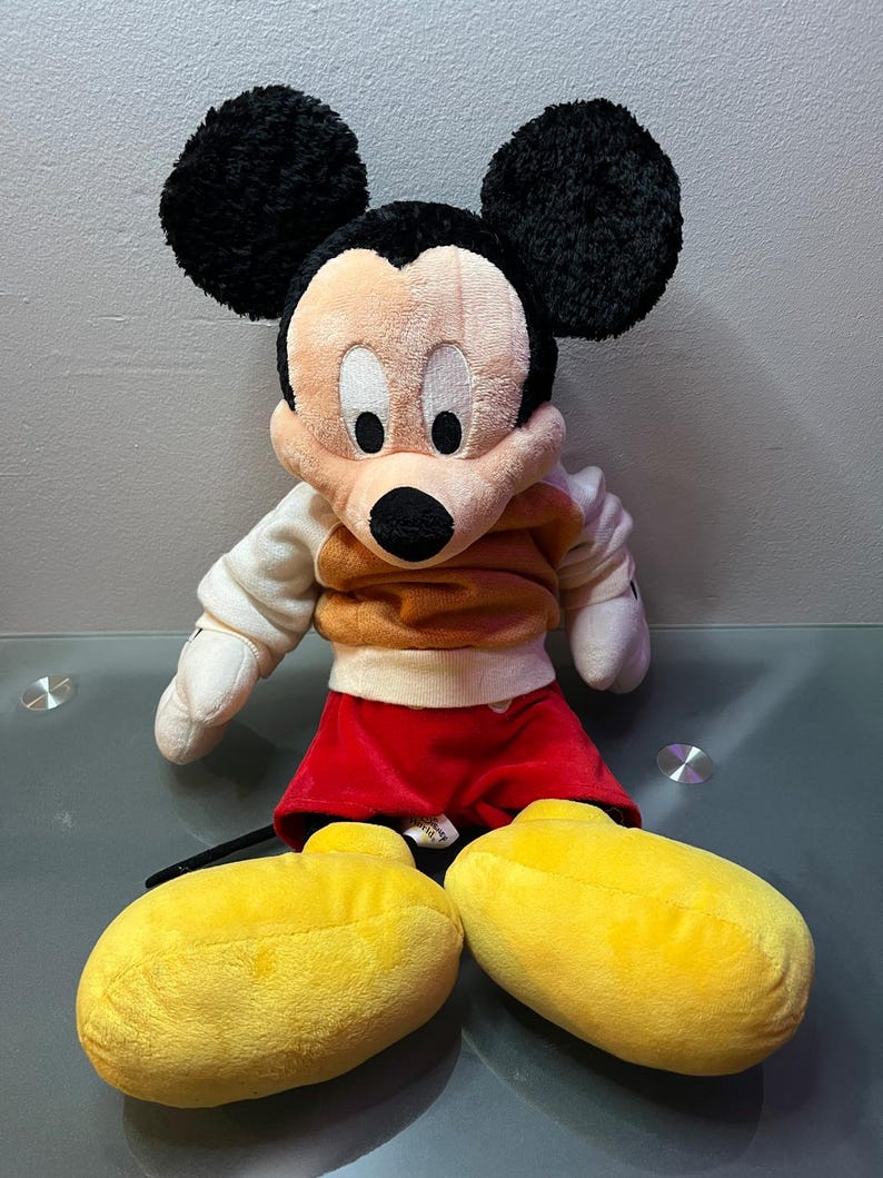 May include: A plush Mickey Mouse doll with black ears, a tan face, and a black nose. He wears a tan and orange shirt, red shorts, and large yellow shoes. The doll is a classic Disney character.
