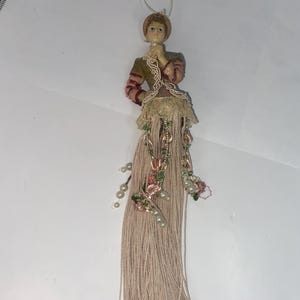 Puede incluir: Un adorno de borla decorativo con una figura de una mujer con un vestido vintage. La figura lleva una chaqueta marrón y roja con un cuello de encaje. La borla es de color marrón claro y está adornada con flores rosas y perlas.