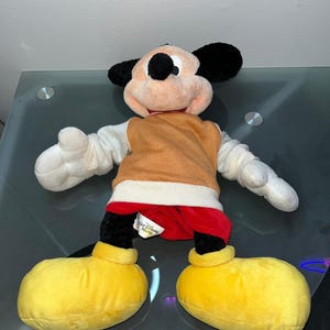 May include: A plush Mickey Mouse doll with black ears, a peach-colored face, and a brown vest. The doll has white gloves, red shorts, and large yellow shoes. A tag reads "Walt Disney World".