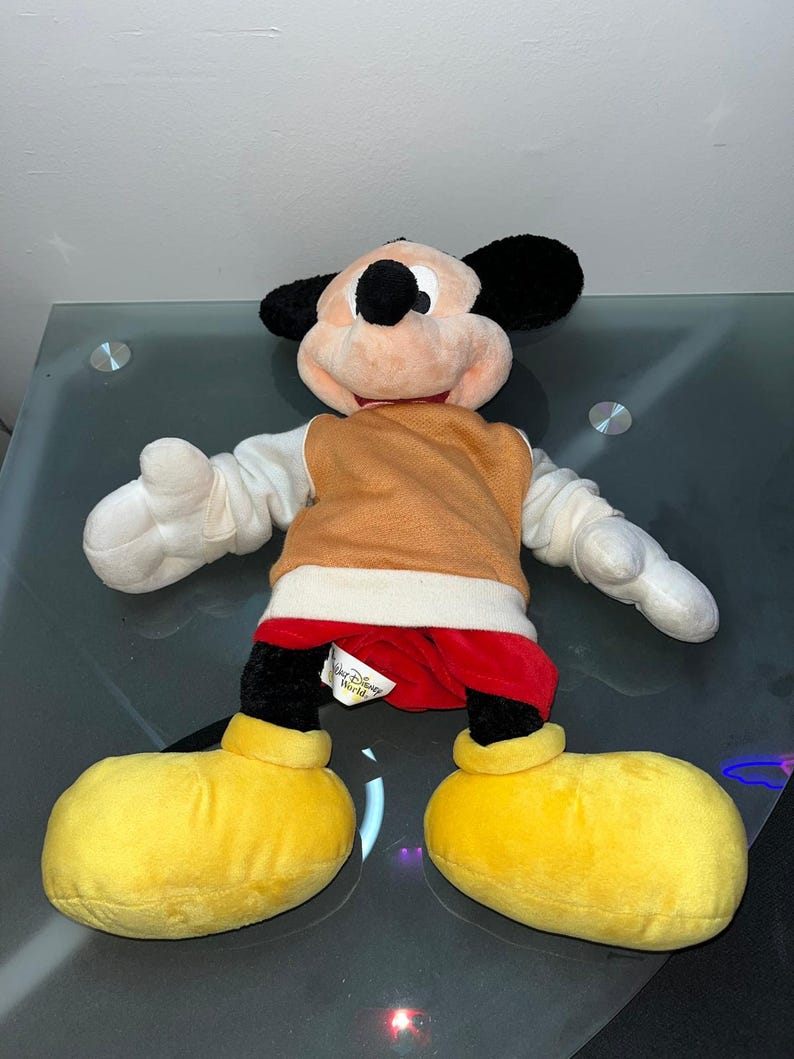 May include: A plush Mickey Mouse doll with black ears, a peach-colored face, and a brown vest. The doll has white gloves, red shorts, and large yellow shoes. A tag reads "Walt Disney World".