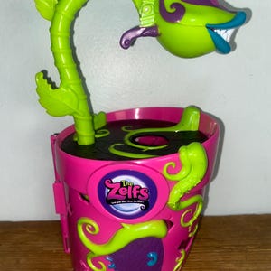May include: A pink plastic toy flower pot with a green, purple, and blue plant-like figure. The pot has a door and the text "The Zelfs". The plant has a smiling face and a curved stem with leaves.