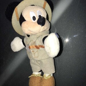 May include: A plush Mickey Mouse toy dressed in a safari outfit. The toy has a tan safari hat, vest, and shorts. The shoes are brown, and the gloves are white. The Mickey Mouse toy is a collectible item.