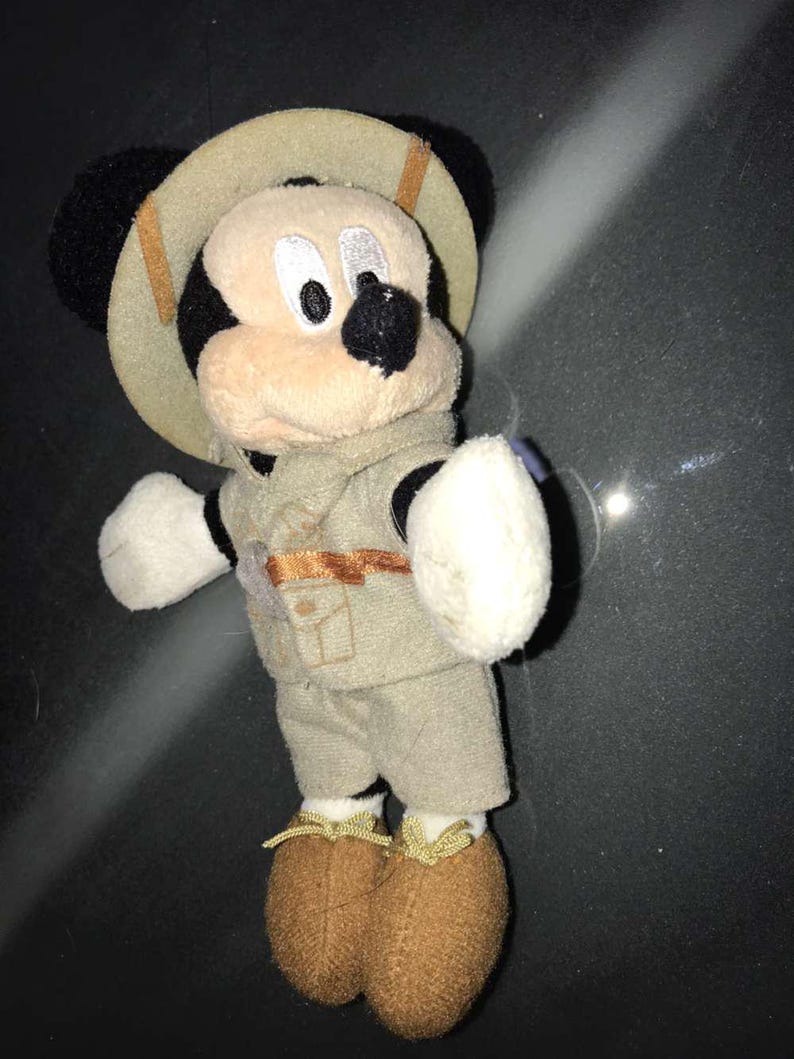 May include: A plush Mickey Mouse toy dressed in a safari outfit. The toy has a tan safari hat, vest, and shorts. The shoes are brown, and the gloves are white. The Mickey Mouse toy is a collectible item.