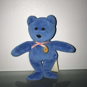 May include: A blue plush teddy bear with a pink ribbon around its neck. The bear has black button eyes and a small patch on its chest. A tag is attached to the bear's side.