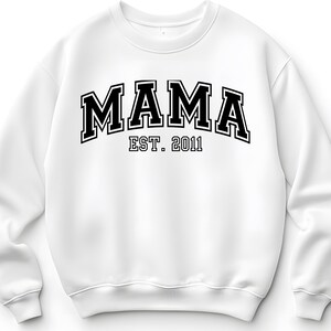 May include: White sweatshirt with the word "MAMA" in bold, black, collegiate-style lettering. Below the word "MAMA" is the text "EST. 2011" in a smaller, black font. The sweatshirt has a crew neck and long sleeves.