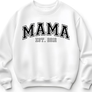 Pode incluir: Camisola branca com a palavra "MAMA" em letras pretas e em negrito, estilo universitário. Abaixo, o texto "EST. 2012" está escrito em preto e em tamanho menor. A camisola tem gola redonda e mangas compridas.
