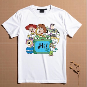 May include: White t-shirt featuring a colorful cartoon graphic of characters, including Woody, Buzz Lightyear, and Jessie. A green frog-like character holds a sign that says "Hi!" in black letters. The shirt has a crew neck.