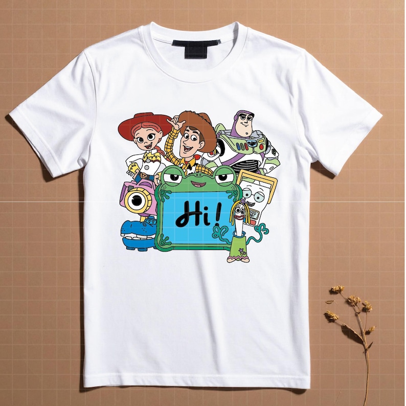 May include: White t-shirt featuring a colorful cartoon graphic of characters, including Woody, Buzz Lightyear, and Jessie. A green frog-like character holds a sign that says "Hi!" in black letters. The shirt has a crew neck.