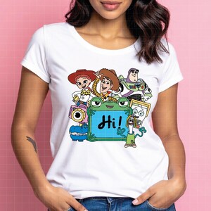 May include: White t-shirt featuring a colorful graphic of characters from the animated film Toy Story. The design includes Woody, Buzz Lightyear, Jessie, and others, with the word "Hi!" on a green sign.