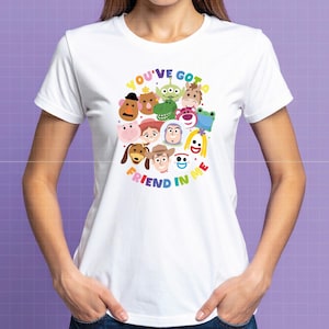 May include: A white short-sleeved t-shirt featuring a colorful graphic print of various animated toy characters arranged in a circular pattern. The text "YOU'VE GOT A FRIEND IN ME" is printed in multicolored letters around the central character cluster.