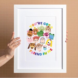 May include: A white framed print featuring a colorful illustration of various cartoon characters arranged in a circular pattern. The text "YOU'VE GOT A FRIEND IN ME" is written in multi-colored letters around the character cluster on a white background.