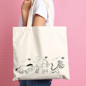 Puede incluir: Bolso tote de color crema con un dibujo lineal en blanco y negro de Winnie the Pooh y sus amigos. El dise&ntilde;o incluye a &Iacute;gor, Piglet, Winnie the Pooh y Tigger, con abejas y l&iacute;neas de puntos. El bolso tiene dos asas color crema.