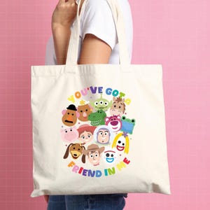 May include: A cream-colored canvas tote bag featuring a colorful graphic print of various animated toy characters arranged in a circular cluster. The text "YOU'VE GOT A FRIEND IN ME" is printed in multi-colored letters arching around the characters.