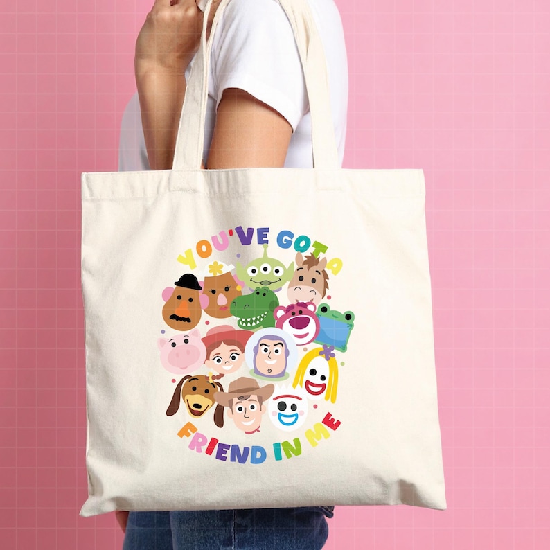 May include: A cream-colored canvas tote bag featuring a colorful graphic print of various animated toy characters arranged in a circular cluster. The text "YOU'VE GOT A FRIEND IN ME" is printed in multi-colored letters arching around the characters.