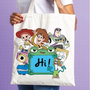 May include: A white tote bag featuring a colorful graphic of animated characters, including a cowboy, a space ranger, and a cowgirl. A green frog-shaped frame with the word "Hi!" in black script is in the center. The bag is held by a person.