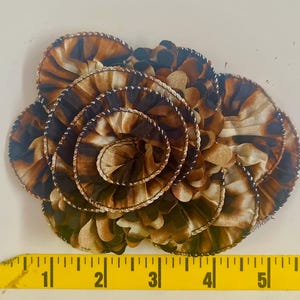 May include: A fabric flower with a brown and tan animal print design. The flower is made of layered, circular fabric pieces with a spiral center. The edges are trimmed with a silver thread. A yellow measuring tape is at the bottom.