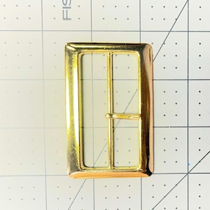 May include: A rectangular gold-colored belt buckle with a single bar and a prong. The buckle has a shiny finish and is designed for use with belts or straps. The buckle is made of metal.