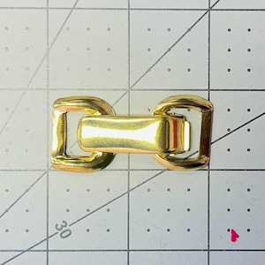 May include: A gold-colored metal clasp, featuring a rectangular center piece connecting two square-shaped loops. The clasp is designed with rounded edges and a shiny finish, suitable for various crafting projects.