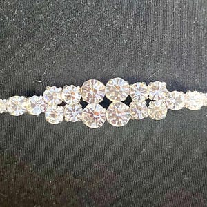 May include: A silver-toned hair clip adorned with multiple clear, sparkling rhinestones. The rhinestones are arranged in a linear pattern, with larger stones clustered in the center and smaller ones on the ends. The clip is set against a dark background.