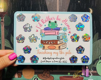 May include: A light blue sticker with the text "I'd Rather be Home" and "Finishing my tbr pile". The sticker features a stack of books with a frog on top, surrounded by holographic flower stickers.
