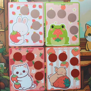May include: Four colorful scratch-off cards with cute illustrations. A bunny, frog, cat, and pig each hold strawberries. Each card has brown circles for scratching. The cards are decorated with strawberries and the text "@budgetinginafairytale".