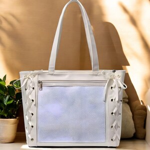 Puede incluir: Bolso tote blanco con un panel frontal de malla rectangular y cierre de cremallera. Los lados presentan un diseño de cordones con cinta blanca. El bolso tiene dos asas largas y está hecho de un material liso similar al cuero.