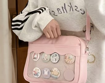 May include: A light pink crossbody bag with a clear front panel displaying several round, colorful pins with cartoon designs. The bag has a matching pink strap and silver hardware. The person is wearing a white sweatshirt.