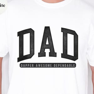 May include: White t-shirt with the word "DAD" in large, bold, black letters. Below the word "DAD" is the phrase "DAPPER AWESOME DEPENDABLE" in a smaller, black font. The shirt is a crew neck style.