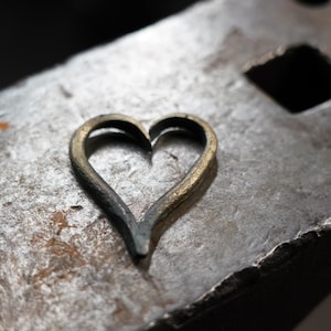 May include: A handcrafted metal heart shape, possibly a pendant or charm, with a textured, aged appearance. The heart is set against a rough, metallic surface, suggesting a blacksmithing or industrial theme.