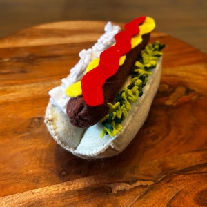 Puede incluir: Un perrito caliente de fieltro hecho a mano con una salchicha marrón, ketchup rojo, mostaza amarilla y relish verde, metido en un panecillo blanco. El perrito caliente está sobre una superficie de madera.