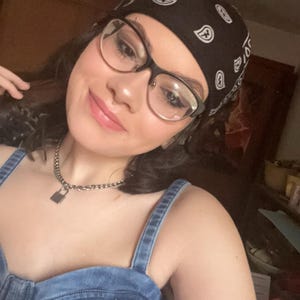 Puede incluir: Un pañuelo negro con un estampado de cachemira blanco se lleva en la cabeza. La persona lleva gafas con montura negra y una parte superior de mezclilla. También es visible un collar de cadena plateada con un colgante de candado.