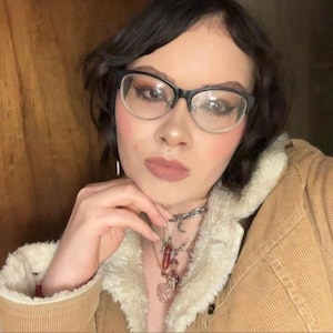 May include: A person wearing black-framed glasses, a corduroy jacket with a cream-colored shearling collar, and a silver necklace with multiple pendants. The person has dark hair and is looking at the camera.