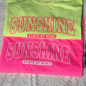 May include: Two t-shirts stacked on top of each other. The top shirt is neon yellow with the words "Sunshine State of Mind" in pink lettering. The bottom shirt is hot pink with the same words in white lettering.