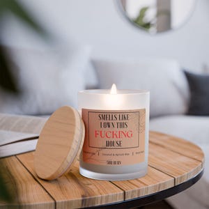 May include: A frosted glass candle with a wooden lid. The candle is lit and the label reads "Smells like I own this FUCKING HOUSE". The candle is on a wooden table.