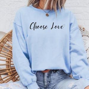 以下が含まれることがあります： 薄い青色のスウェットシャツに、黒い筆記体で「Choose Love」という文字と小さなハートのマークが書かれています。