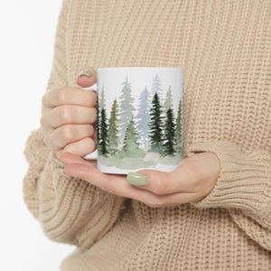 May include: White ceramic mug featuring a watercolor design of evergreen trees in shades of green and blue. The mug has a standard handle and is held by a person wearing a beige sweater. The design wraps around the mug, creating a forest scene.
