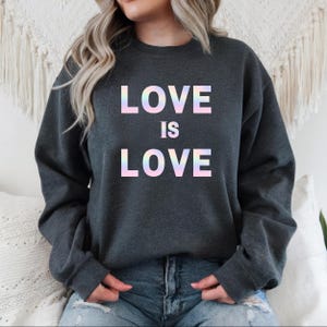 Op de afbeelding: Een donkergrijs sweatshirt met de tekst "LOVE IS LOVE" gedrukt in regenboogkleurige, iriserende letters.