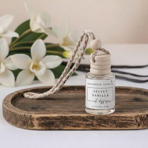 Puede incluir: Un difusor de aceite de vidrio transparente etiquetado "Velvet Vanilla" con una tapa de madera y un cordón tejido. El difusor se encuentra sobre una bandeja de madera rústica, con flores blancas y vainas de vainilla en el fondo. Contiene 8 ml.