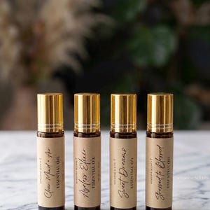 Könnte beinhalten: Vier bernsteinfarbene Glasfläschchen für ätherische Öle mit goldenen Verschlüssen und beigen Papieretiketten. Die Etiketten tragen den schwarzen Text Clear Mind + Air, Ageless Elixir, Sweet Dreams und Stressed to Blessed sowie den Markennamen.
