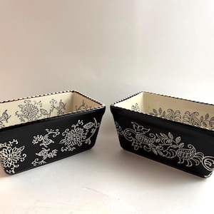 May include: Two rectangular black ceramic baking dishes with white floral designs. The interior is cream-colored with a white floral pattern. The rims are decorated with a dotted pattern. The dishes are ideal for baking.