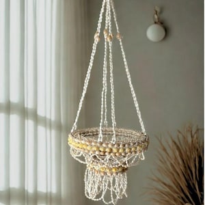 May include: A hanging plant holder made of off-white macrame cord and adorned with seashells and wooden beads. The holder has a circular basket shape with a fringed bottom, designed to suspend a potted plant. The background includes a window and a neutral-colored wall.